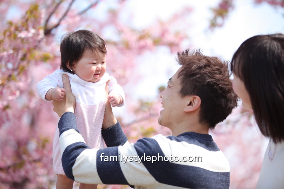 桜を背景に赤ちゃんを高い高いするパパとそれを見つめるママ